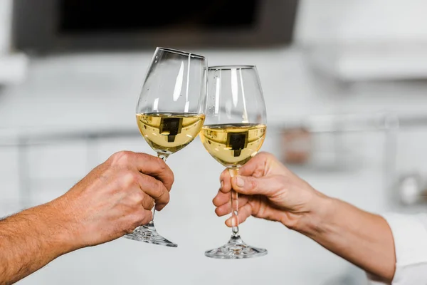 Imagen recortada de la esposa y el marido tintineo con vasos de vino blanco en la cocina - foto de stock