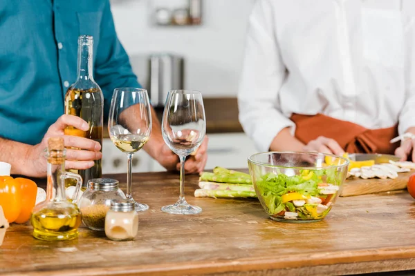 Cropped image of mature wife cutting vegetables and husband holding bottle of wine in kitchen — Stock Photo