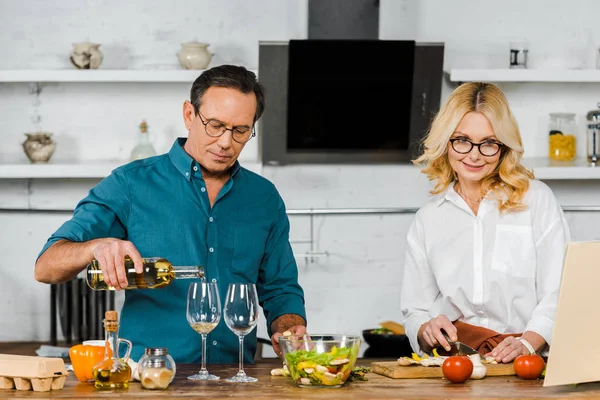 Bonito maduro marido derramando vinho em copos, atraente esposa cortando legumes na cozinha — Fotografia de Stock