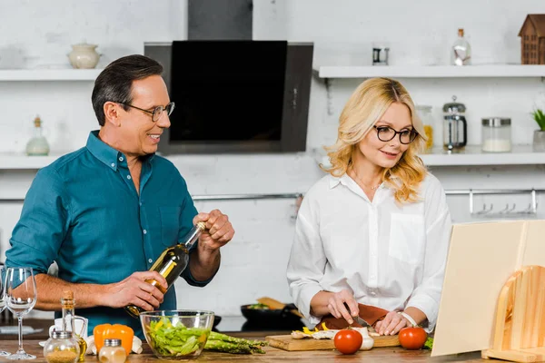 Reife Frau schneidet Gemüse und Ehemann hält Flasche Wein in der Küche — Stockfoto