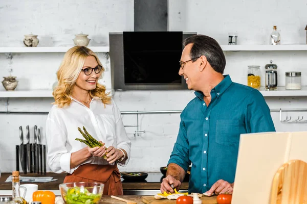 Happy mature wife and husband cooking together and looking at camera in kitchen — Stock Photo