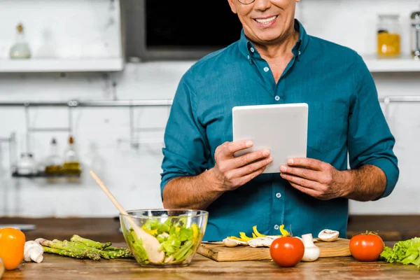 Abgeschnittenes Bild eines lächelnden reifen Mannes mit Tablette beim Kochen in der Küche — Stockfoto