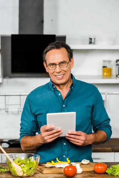 Smiling handsome mature man using tablet while cooking in kitchen, looking at camera — Stock Photo