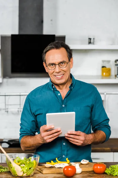 Sonriente guapo maduro hombre utilizando tableta mientras cocina en cocina, mirando a la cámara - foto de stock