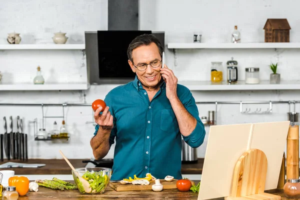 Handsome mature man talking by smartphone while cooking in kitchen — Stock Photo