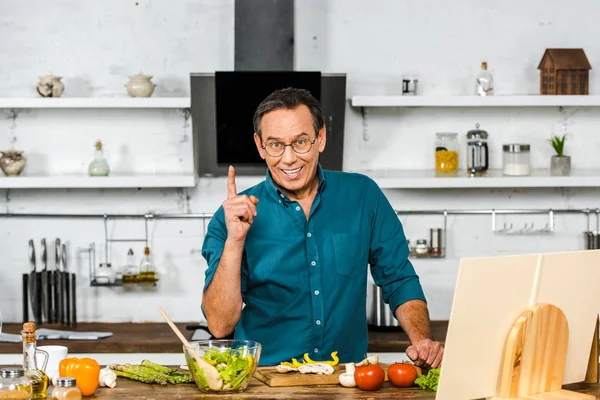 Sonriente guapo maduro hombre cocina en cocina y mostrando un dedo arriba - foto de stock