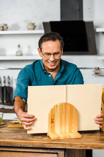 Smiling handsome mature man cooking in kitchen and taking cookbook — Stock Photo