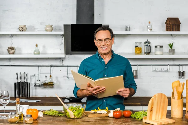 Handsome mature man cooking in kitchen and holding cookbook — Stock Photo