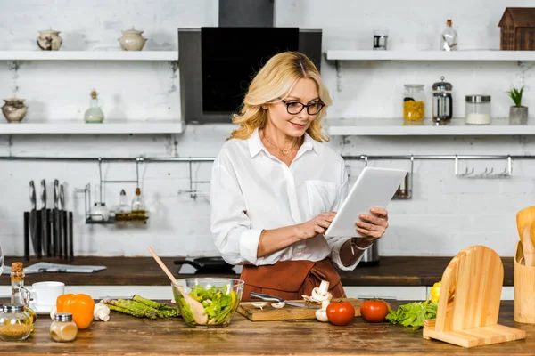 Atractiva mujer madura en gafas de lectura receta de la tableta durante la cocción en la cocina - foto de stock