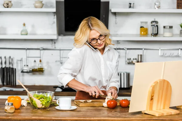 Mulher madura atraente falando por smartphone, cortando legumes e olhando para livro de receitas na cozinha — Fotografia de Stock