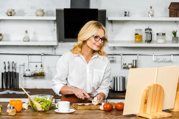 Attractive mature woman looking at recipe book while cutting mushrooms in kitchen — Stock Photo