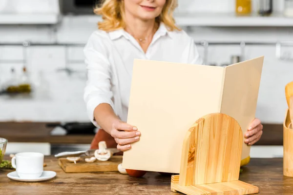 Immagine ritagliata di donna matura che prende libro di ricette per cucinare in cucina — Foto stock