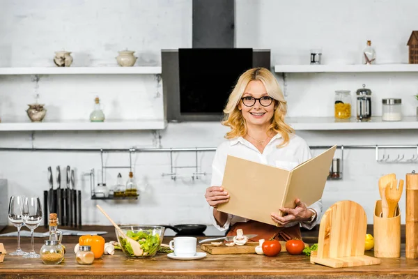 Усміхнена приваблива зріла жінка тримає книгу рецептів для приготування їжі на кухні і дивиться на камеру — стокове фото
