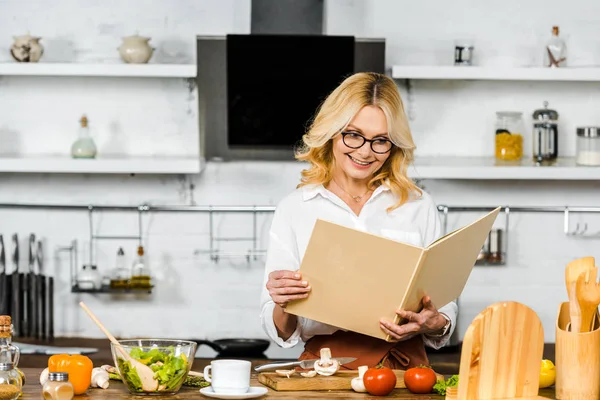 Улыбающаяся привлекательная зрелая женщина читает рецепт книги для приготовления пищи на кухне — стоковое фото