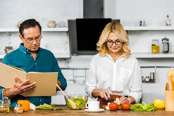 Esposa madura y marido cocinando junto con el libro de recetas en la cocina - foto de stock