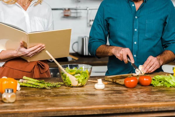 Cropped image of mature wife and husband cooking together with recipe book in kitchen — Stock Photo