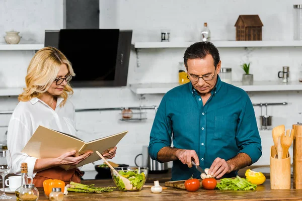 Frau und Mann mittleren Alters kochen zusammen mit einem Rezeptbuch in der Küche — Stockfoto