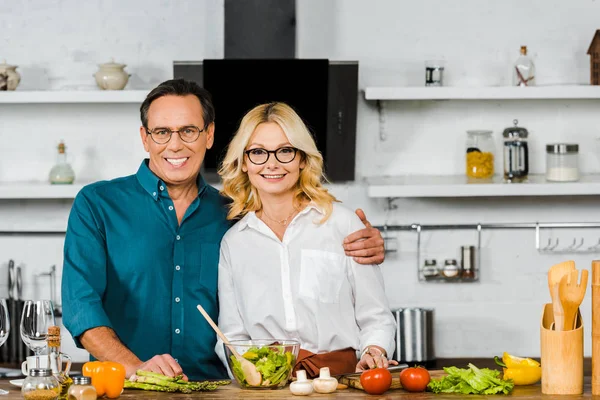 Sonriente madura esposa y marido abrazando y mirando a la cámara en la cocina — Stock Photo