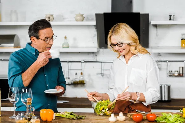 Attractive mature wife mixing salad and husband drinking tea in kitchen — Stock Photo