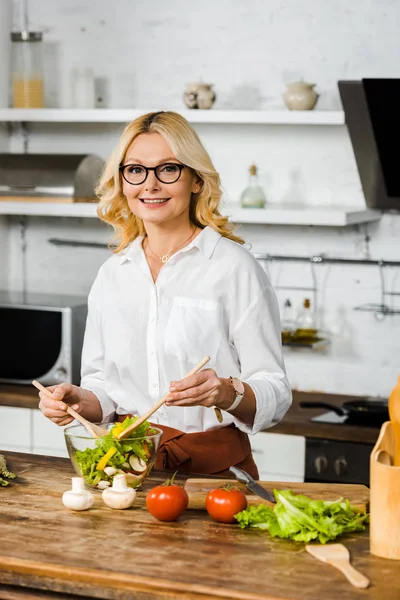 Sonriente hermosa mujer de mediana edad mezclando ensalada en cuenco de vidrio en la cocina y mirando a la cámara - foto de stock