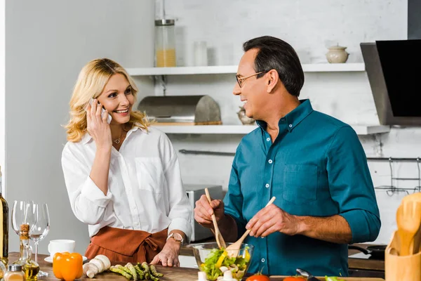 Esposa madura hablando por teléfono inteligente y marido mezcla ensalada en un tazón en la cocina - foto de stock