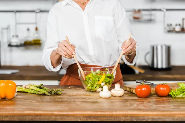 Обрезанный образ зрелой женщины, смешивающей салат в стеклянной миске на кухне — стоковое фото