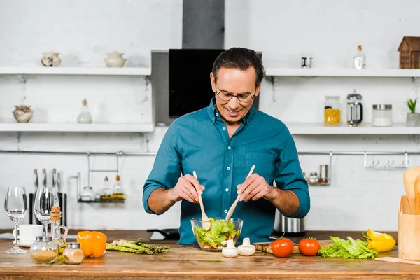 Bel homme mature dans des lunettes mélangeant la salade dans un bol dans la cuisine — Photo de stock