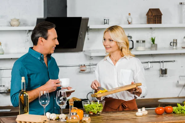 Lächelnd attraktive reife Frau legt Gemüse in Schüssel und Ehemann hält Tasse Tee in der Küche — Stockfoto