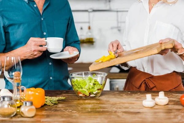 Image recadrée de femme mature mettant des légumes dans un bol et mari tenant une tasse de thé dans la cuisine — Photo de stock