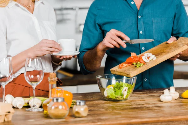 Imagen recortada de marido maduro poniendo verduras en tazón y esposa sosteniendo taza de té en la cocina - foto de stock