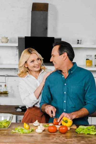 Sonriendo esposa madura abrazando marido mientras él cocina ensalada en la cocina, mirándose el uno al otro - foto de stock