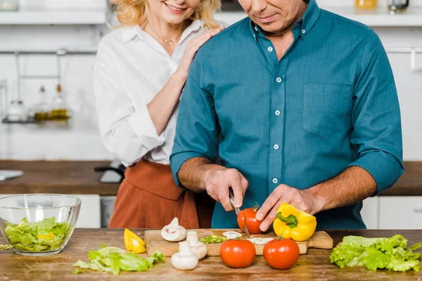Cropped image of mature wife hugging husband while he cooking salad in kitchen — Stock Photo