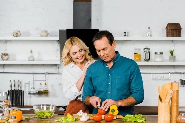 Esposa madura abrazando marido guapo mientras él cocina ensalada en la cocina - foto de stock