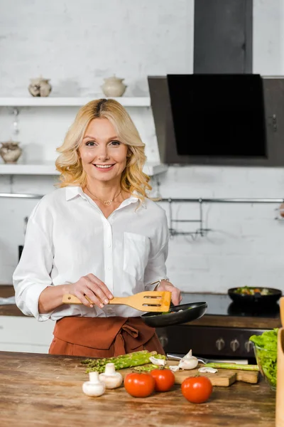 Sonriente atractiva mujer de mediana edad sosteniendo sartén para preparar verduras en la cocina - foto de stock