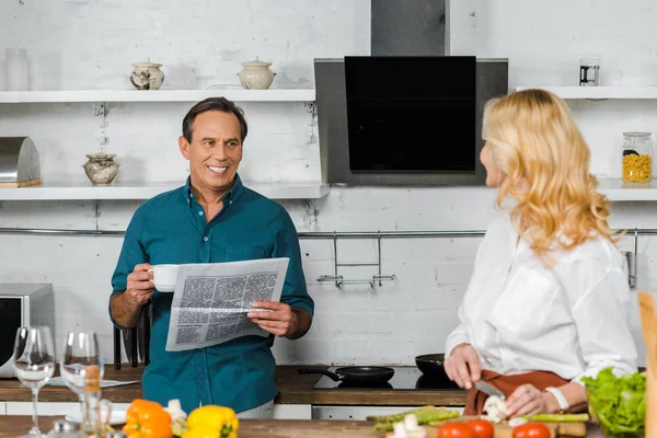 Mature wife cooking and smiling husband reading newspaper in kitchen — Stock Photo