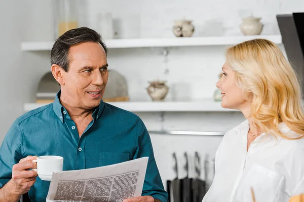 Mature wife looking at husband in kitchen, he holding cup of tea and newspaper — Stock Photo