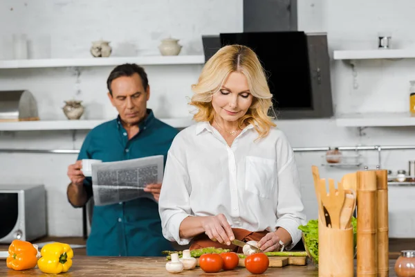 Moglie matura che taglia verdure e marito che legge giornale in cucina — Foto stock