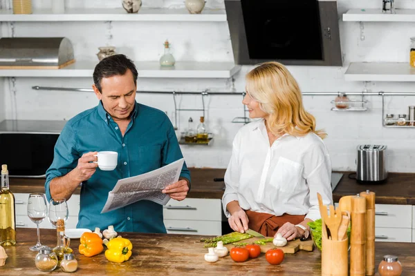 Attraktive reife Frau schneidet Gemüse und Mann liest Zeitung in der Küche — Stockfoto