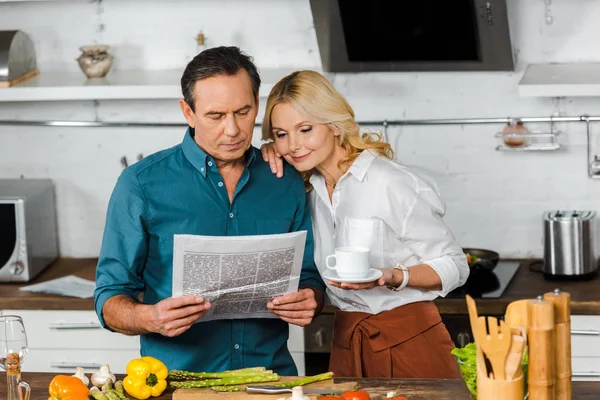 Зрелая жена держит чашку чая и муж читает газету на кухне — стоковое фото