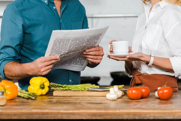 Immagine ritagliata di moglie matura che tiene una tazza di tè e marito che legge il giornale mentre cucina in cucina — Foto stock
