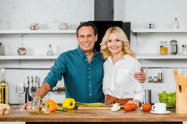 Lächelnd reife Frau und Mann umarmen sich und schauen in die Kamera am Tisch in der Küche — Stockfoto