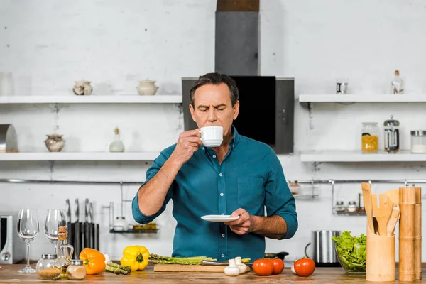 Guapo hombre de mediana edad bebiendo té con los ojos cerrados mientras cocina en la cocina - foto de stock