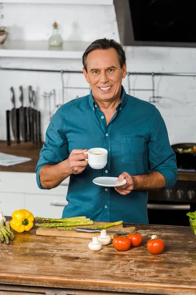 Guapo sonriente hombre de mediana edad sosteniendo la taza de café y mirando a la cámara en la cocina - foto de stock