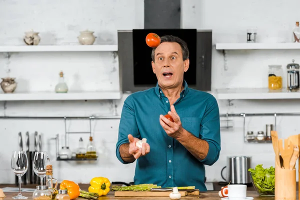 Sorprendido guapo de mediana edad hombre malabares verduras mientras cocina en la cocina - foto de stock