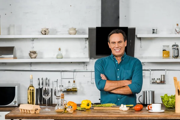 Lächelnder schöner Mann mittleren Alters, der mit verschränkten Armen in der Küche steht und in die Kamera schaut — Stockfoto