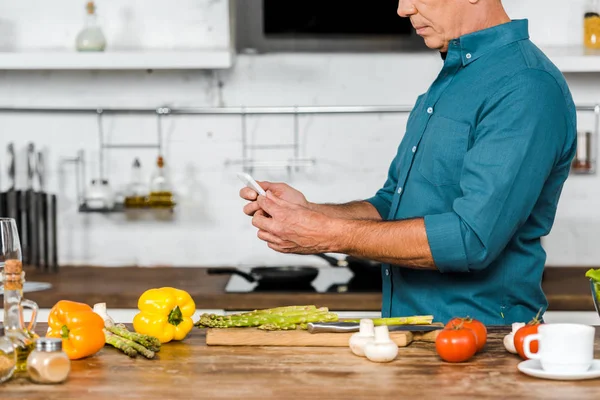 Immagine ritagliata di uomo di mezza età utilizzando smartphone durante la cottura in cucina — Foto stock