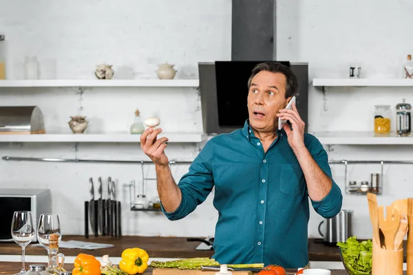 Guapo hombre de mediana edad hablando por teléfono inteligente mientras cocina en la cocina - foto de stock