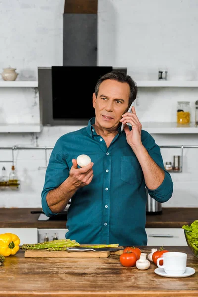 Handsome middle aged man talking by smartphone and holding mushroom in kitchen — Stock Photo