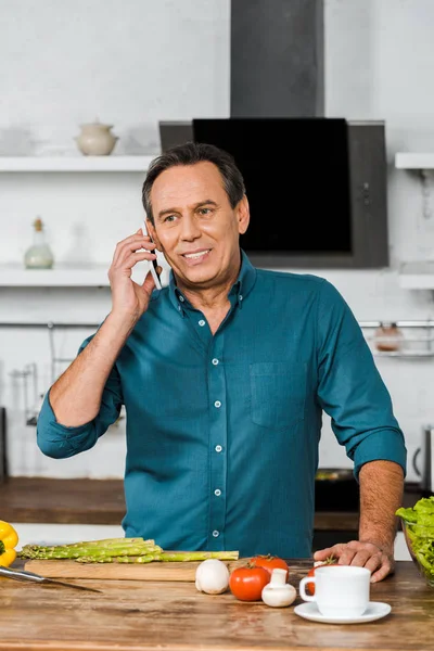 Handsome mature man talking by smartphone while cooking in kitchen — Stock Photo