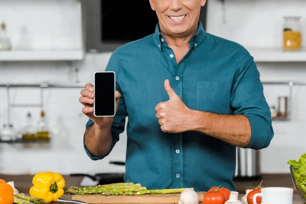 Abgeschnittenes Bild eines lächelnden gutaussehenden Mannes mittleren Alters, der ein Smartphone mit leerem Bildschirm hält und den Daumen in der Küche nach oben zeigt — Stockfoto